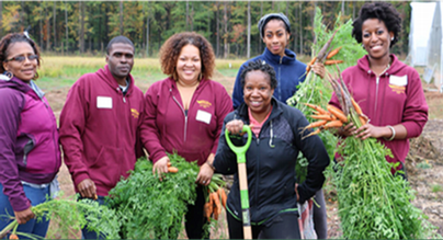 A group of people smiling while holding carrots and shovels in a garden setting.