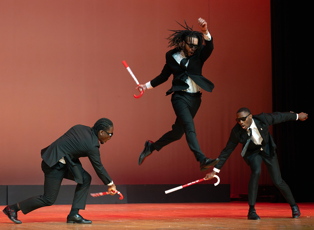 Men on a stage dressed in black suits holding canes. One jumping in the air.
