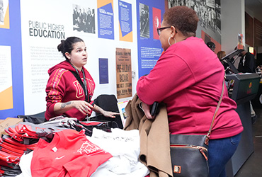 Woman standing a table of tshirts talking to another woman on the other side of the table.