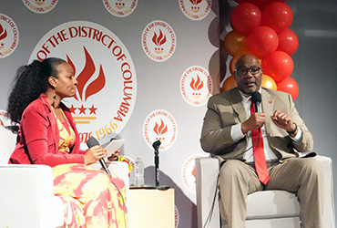 A woman and a man sitting on chairs on a stage having a public discussion.